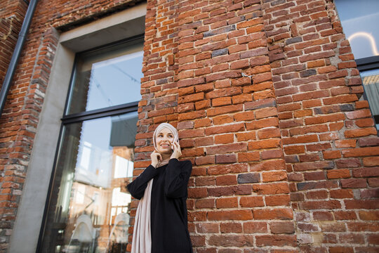 Cheerful young islamic businesswoman talking on mobile phone while standing near red brick wall. Beautiful young woman with hijab working using smartphone during break.