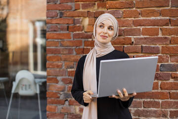 Portrait of smiling muslim woman relaxing using digital laptop while standing near red brick wall. Young muslim girl looking at screen of notebook speaking during online video call on social media.