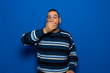 Hispanic man wearing sweater standing over isolated blue background surprised covering mouth with hand for mistake. Secret concept