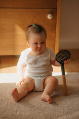 A 7-month girl is sitting on the carpet dressed in a bodysuit at home. A cute infant is playing...