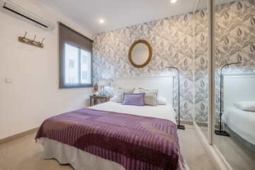 Fototapeta na wymiar Bedroom with double bed, built-in wardrobe with sliding mirror doors and white wooden headboard, air conditioning, walls with decorative paper and window with blinds.