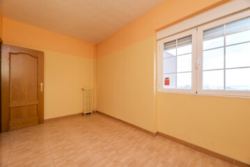 Empty room with light brown stoneware floor, bicolor yellow painted walls and white aluminum windows, cast iron radiator and oak wood door