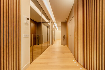 Hallway of a house with a wall covered with mirrors on one side and wooden slats on the other, chestnut floorboards and recessed lights in the floor