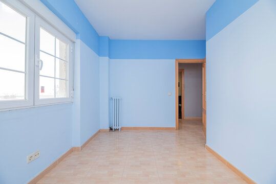 Empty room with light brown stoneware flooring, cast iron radiator and two tone blue painted walls and white aluminum windows
