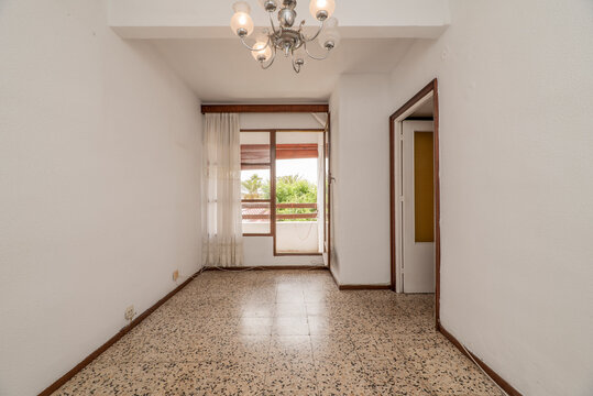 Empty room with old terrazzo floor, white walls and windows with access to a terrace with white curtains, wooden railings and views of a garden with trees