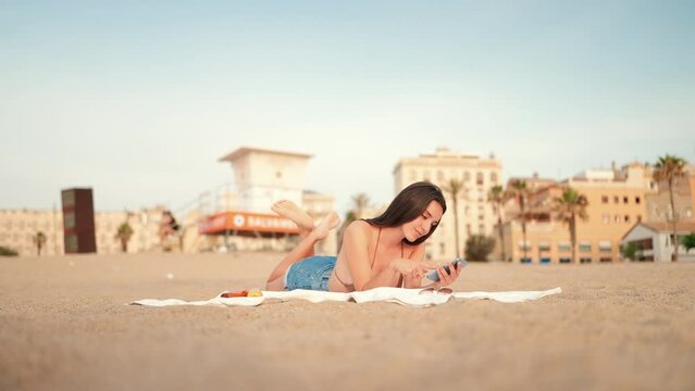 Slim beautiful brown-haired woman with long hair uses mobile phone while relaxing on the beach. Smiling girl wearing swimsuit and denim shorts scrolling information on smartphone. slow motion