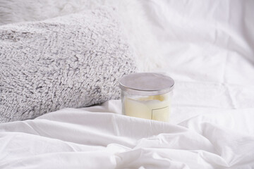 Blank off-white pillar candle in glass jar with label and silver colored lid on white bed sheets...