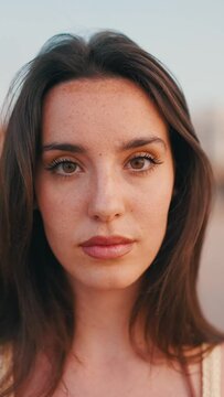 VERTICAL VIDEO: Close-up Beautiful brown-haired woman with long hair sits on the beach. Gorgeous girl with freckles and long eyelashes looks at the camera. slow motion