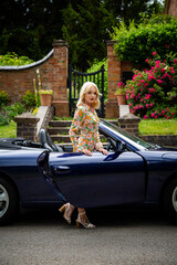 Beautiful young woman standing within the door of a blue Porsche sports car with a garden gate and...
