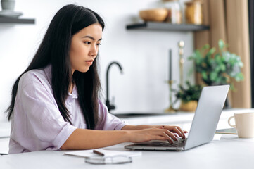 Thoughtful smart young chinese woman, sit at home in the kitchen, working or study online, focused looks at laptop screen, planning and analysis future project, startup, female student doing homework