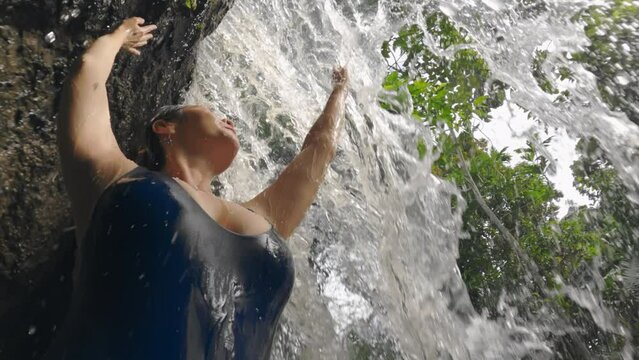 A girl with big breasts in blue swimsuit touching powerful waterfall stream on tropical. Caucasian sexy girl stands under a rock from which a stream of mountain river flows on sunny day. Slow motion