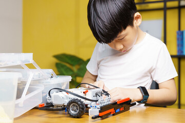 Smart looking Asian student kid assembling robot, coding and solving engineering problem at home with yellow wall in background. STEM education and 21st century learning skills concept