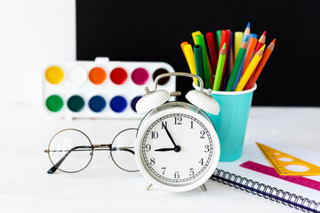 Time to return back to school. Bright colourful green yellow, blue stationery, paints, pencils, markers, brushes. black board on background. White alarm clock. Copy space for text. Autumn, September