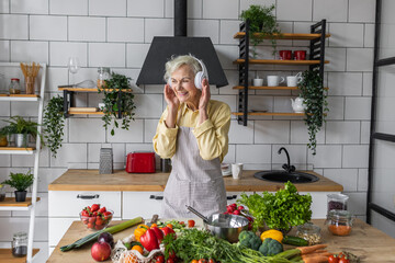 Senior attractive woman listening to music and singing in her headphones in the kitchen. Active lifestyle of mature people, enjoying life, having fun. Using whisk as microphone while cooking food