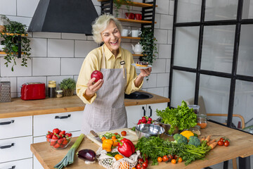 Attractive smiling senior woman with apple and donut, looking happy and healthy. Concept of active lifestyle of mature people, organic food. Female at home preparing vegetarian salad, holding a cake