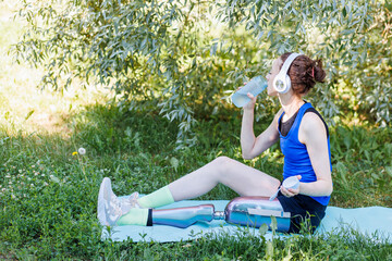Young beautiful athletic woman with bionic prosthetic leg doing sport exercises in the park outdoors in summer. Concept of active lifestyle of disabled people. Listening to music, drinking water