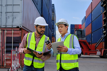 Two professional Asian male workers in safety uniforms and hard hats work at a logistics terminal with many stacks of containers, loading control shipping goods for the cargo transportation industry.