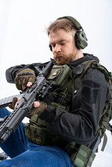 Fototapeta premium airsoft player in full gear with GG RK74 fire series guns. a man in headphones, a bulletproof vest, with a backpack and a belt, aims his machine gun to the side. profile. White background.