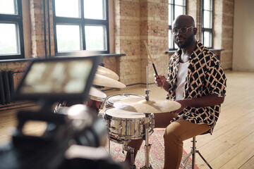 Portrait of African young drummer performing on drums on camera for online broadcasting at empty...