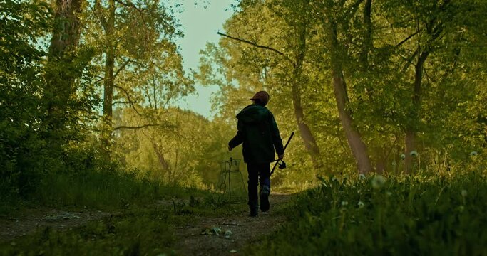 schoolboy with fishing rod and fish trap is going on path in beautiful forest, rear view, 4K, Prores