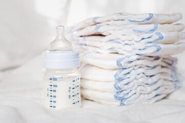 Baby bottle with milk and diapers on a white fabric background. Baby care. First days of life. Feeding and motherhood - obrazy, fototapety, plakaty