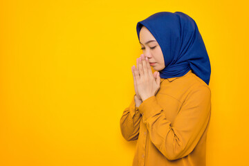 Hopeful young Asian Muslim woman dressed in orange keeps palms pressed together in praying gesture isolated on yellow background