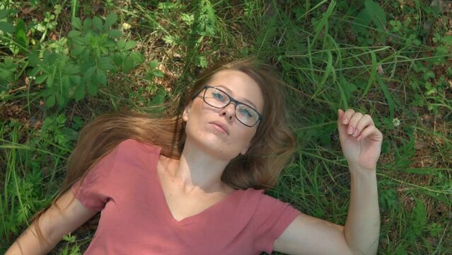 Woman enjoying summer day lying on a grass and dreaming in a forest park. The face of a calm, relaxed and thoughtful girl with glasses in deep thinking. Sunny day woods holiday.