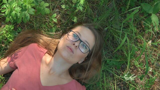 Woman enjoying summer day lying on a grass and dreaming in a forest park. The face of a calm, relaxed and thoughtful girl with glasses in deep thinking. Sunny day woods holiday.
