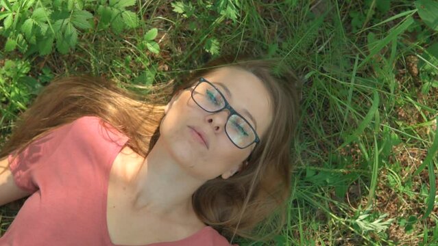 Woman lying on a grass and dreaming in a summer forest park. The face of a calm, relaxed and thoughtful girl with glasses in deep thinking. Sunny day woods holiday. Dramatic zoom out.