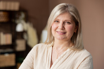 Fototapeta premium Headshot portrait positive mature woman with healthy smile standing on background of cozy living room, happy middle-aged lady looking to camera, posing for photo at home, enjoying leisure time.