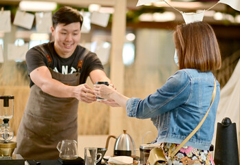 Closeup of Asian Male Barista handing over a cup of coffee to customers with a happy smile and service inside a coffee shop cafe in Thailand.