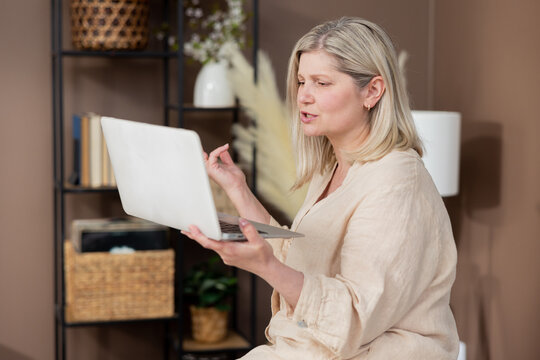 Happy older middle aged mature woman, waving hand gestures holding laptop, video conference, long distance conversation, virtual family chat, online meeting, sitting at home.