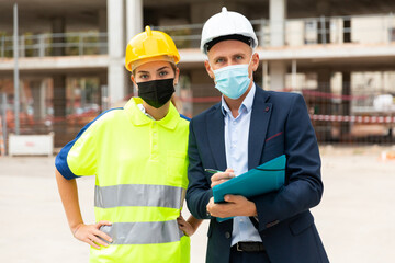 Man architect and a young woman engineer in protective masks, working on a construction site during a pandemic, take ..important notes discussing a construction project