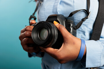 Close up of professional photographer with photo camera getting ready for shooting session. African american young adult woman having modern DSLR device while standing on blue background.