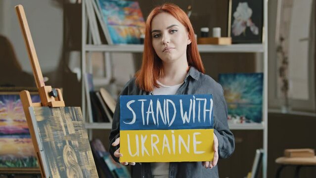 Red-haired talented artist stands posing in art studio workshop against background of drawing picture, painter holds cardboard banner stand with Ukraine support freedom of Ukrainians anti war poster