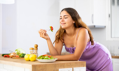 Obraz na płótnie Canvas Portrait of positive young woman in nightie leaning on table, eating salad in kitchen at home. Woman has cooked fresh vegetable salad.