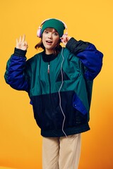 a joyful woman in a green jacket and a hat in the style of the 90s enjoys music standing in pink headphones on a yellow background holding them with her hand and showing a peace sign. Vertical
