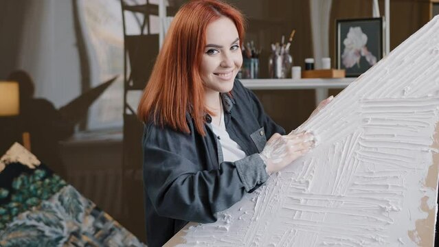 Red-haired 20s Caucasian woman female artist girl painter in workshop draws puts plaster on canvas makes decor picture with hands painting drawing decorating talented art lady looking at camera