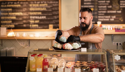 Indian waiter working in a cafe, barista putting meringue cakes on showcase of a coffee shop