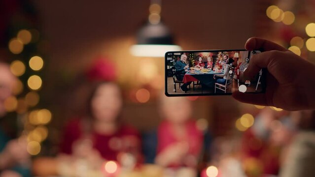 Festive family members getting photographed with smartphone while sitting at table enjoying Christmas dinner . Person taking photo of happy people celebrating winter holiday inside cozy apartment.