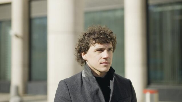 Camera Move Around: A young man with dark curly hair stands, takes out his phone, takes pictures. Against the backdrop of office buildings. 