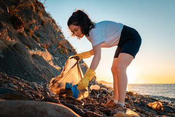 A female volunteer collects plastic bottles in a garbage bag. In the background, the sea and the sunset. The concept of environmental protection and coastal zone cleaning
