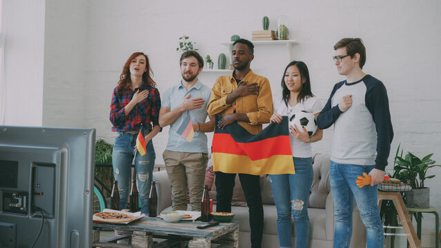 Multi-ethnic group of friends sport fans listening and singing German national anthem before watching sports championship on TV together at home indoors