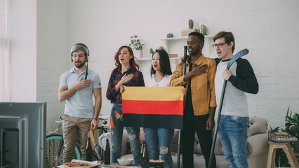 Multi ethnic group of friends sport fans listening and singing German national anthem before watching sports championship on TV together at home indoors - obrazy, fototapety, plakaty