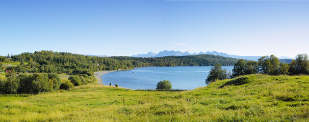 Fototapeta na wymiar Landscape of a lake with trees near a field. Green hills by the seaside with a blue sky in Norway. A calm lagoon near a vibrant wilderness against a bright cloudy horizon. Peaceful wild nature scene