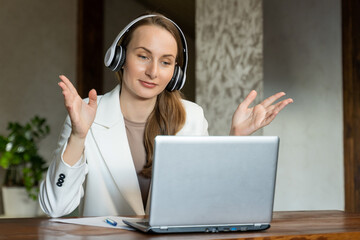 A happy charming young business woman wearing a headset is having a video call on laptop. Young woman in office