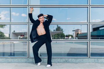 Full body of queer male in black jacket on naked torso standing with raised arm near glass wall in...
