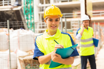 Young attractive girl engineer in hard hat with papers checking work process in construction site outdoors
