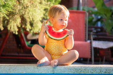 Cute Caucasian little girl with dirty mouth in yellow swimwear funny eating a slice of fresh...