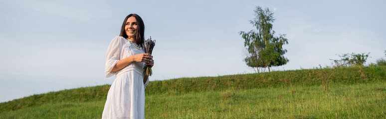 brunette woman in white dress holding lavender bouquet and smiling in green field, banner.